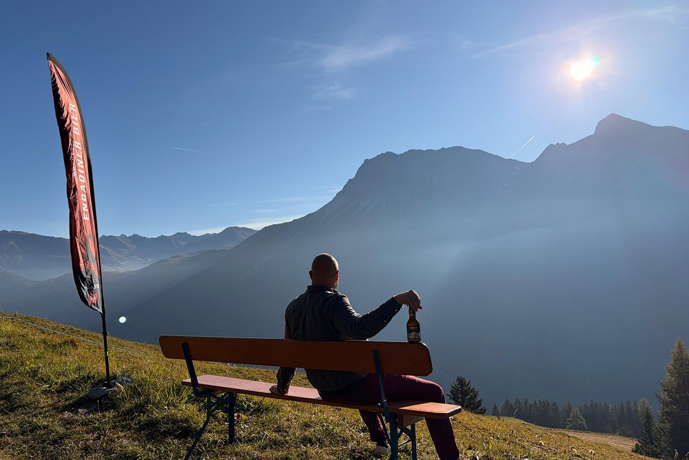 Der Braumeister von Engadiner Bier sitzt am frühen Morgen auf einer Bank über dem Engadin und geniesst den Sonnenaufgang mit einem Bier in der Hand. Neben ihm weht eine Engadiner-Bier-Fahne, während die ersten Sonnenstrahlen die Berggipfel erleuchten.