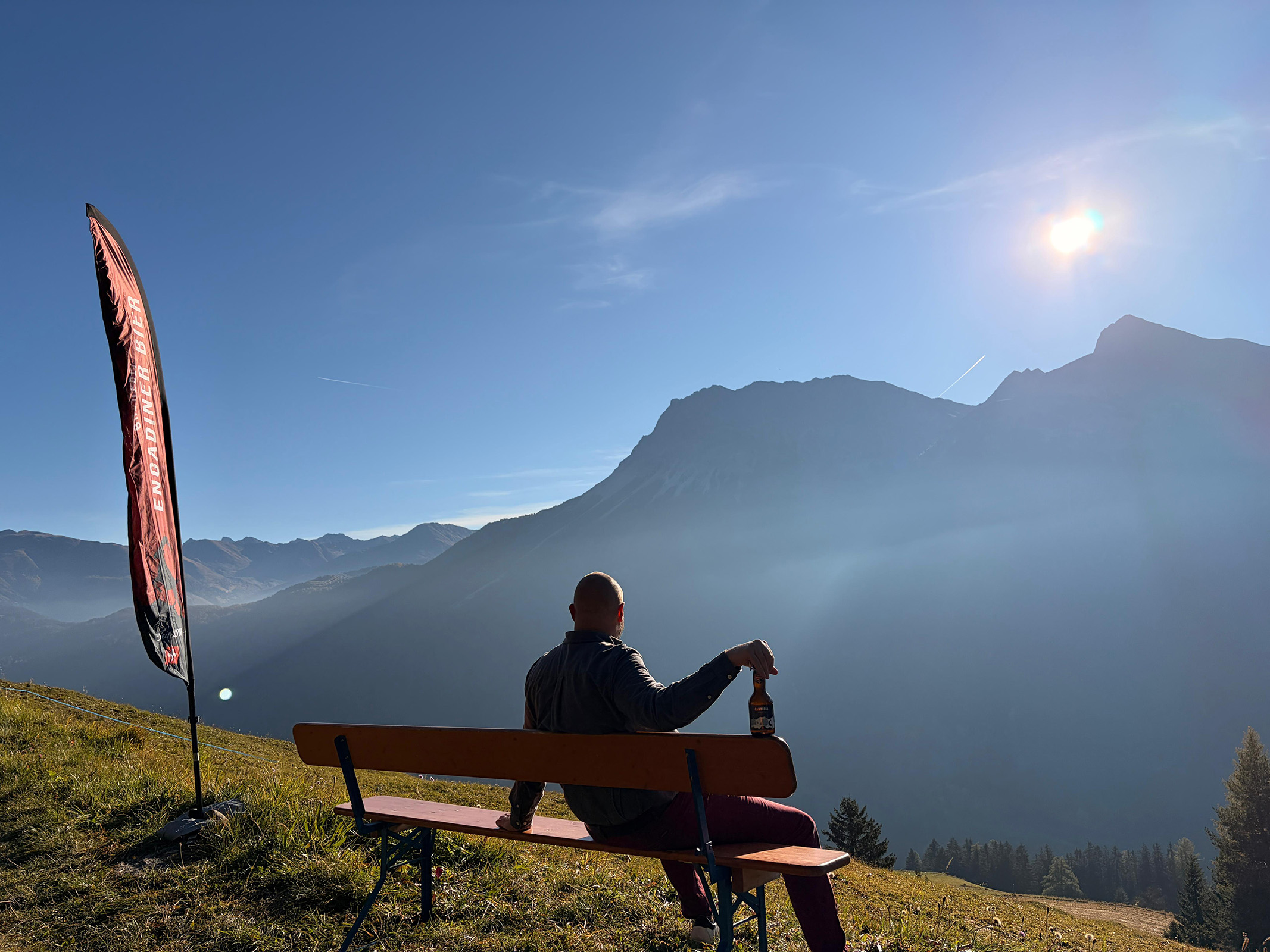Der Braumeister von Engadiner Bier sitzt am frühen Morgen auf einer Bank über dem Engadin und geniesst den Sonnenaufgang mit einem Bier in der Hand. Neben ihm weht eine Engadiner-Bier-Fahne, während die ersten Sonnenstrahlen die Berggipfel erleuchten.